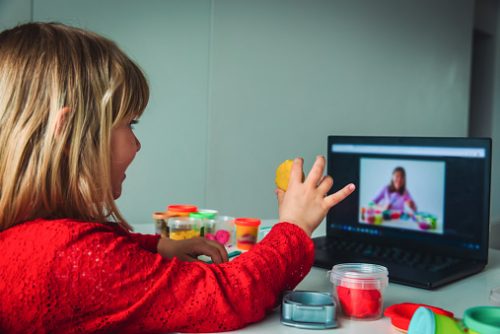 Child in a red shirt participating in online speech therapy in NYC, holding playdough while following a therapist’s activity on a laptop screen.
