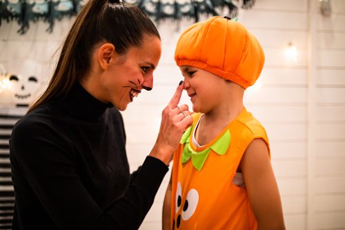 A cute boy in a pumpkin costume and his young mom with cat makeup are smiling while she is bopping the boy's nose