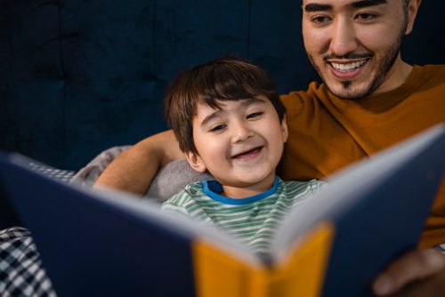 A father and son share a bedtime story, smiling as they read together. Speech therapy in NYC highlights the benefits of storytelling for children’s brain development and speech skills.