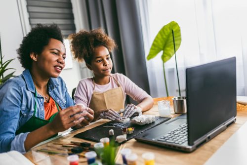 Parent and child engaged in online speech therapy in NYC, working with clay and art supplies while watching instructions on a laptop.