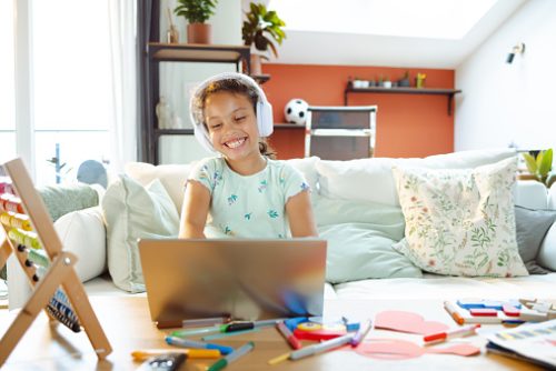 Smiling child wearing headphones during online speech therapy in NYC, surrounded by colorful craft materials and educational toys at home.