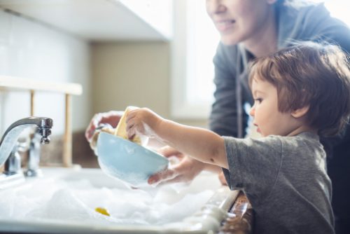 A cute toddler aged boy wearing a blue diaper and tshirt helps out with with washing the dishes, standing on a chair to be able to reach the sink full of soapy bubbles and dishes.  His mother smiles behind him as she supervises his sensory perception experience. Bright sunlight comes in through the window behind him, lighting the sparse modern kitchen.
