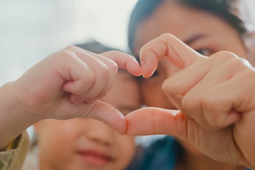 Closeup of young Asian family child making fingers heart shaped signs on sofa in living room at home. Love showing hand heart gesture, Family happy moment concept.
