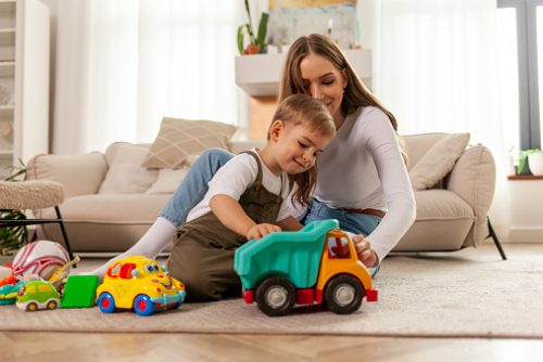 A parent sits on the floor playing with toy vehicles alongside their young child, supporting learning through play as part of early speech therapy goals in NYC.