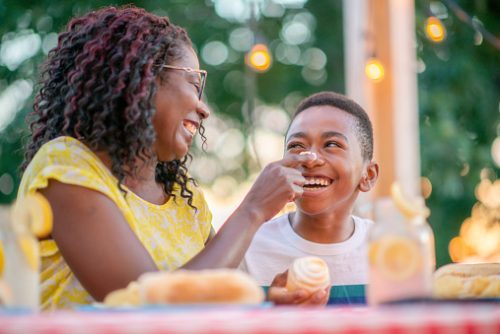 A Mother is seen sitting with her son at a picnic table during a family BBQ.  They are enjoying cold glasses of lemonade and prepared foods, as they laugh and enjoy each others company.