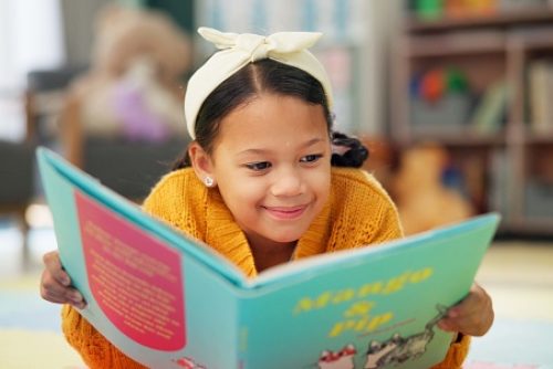 Girl, school and reading book in class, childhood development and growth for vocabulary in kindergarten. Happy, smile and knowledge or information with storybook, learning and fantasy for child