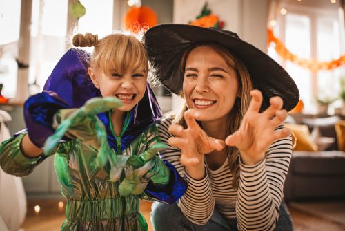 Mother and daughter in costumes decorating and preparing sweet table for Halloween party at home. They are dancing and having fun