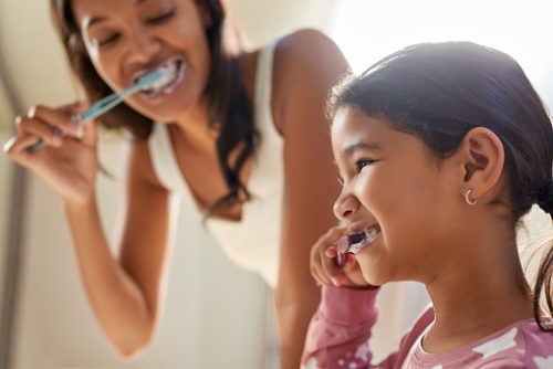 Close up of a smiling daughter and young mother brushing their teeth together with smiles. Cute little girl and her middle eastern mom enjoying a morning routine, brushing their teeth happily.