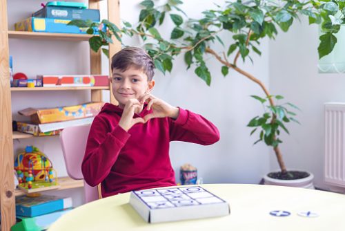 A young autistic boy sitting in a therapy room forming a heart shape with his hands, representing confidence and progress gained through speech therapy for autism in NYC.