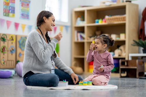 A female Occupational Therapist sits on the floor of a play room with a sweet little mixed race girl, as they learn through play.  The little girl is dressed casually and focused on the toy.