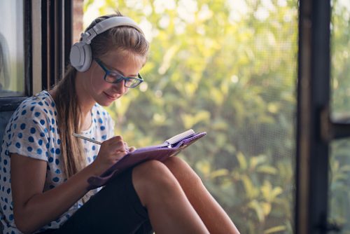 Young girl sitting by a window wearing headphones and glasses, happily writing in a journal, representing self-regulation practices used by a neurodivergent speech therapist in NYC.