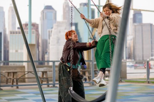 Two European women in their 40s enjoy playground swings against Manhattan skyline backdrop in Brooklyn. Redhead woman in leather jacket pushes her friend who swings joyfully wearing green pants and cream sweater. Both women laugh and smile while reconnecting with childhood fun. Urban recreation area features city views across East River with skyscrapers visible in background. Selective focus on subjects with cityscape providing context.