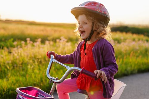A joyous little girl in a helmet laughs exuberantly while sitting on her bicycle against a golden, sunlit field of tall grass.