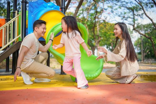 Family playing at a park with a young girl going down a slide while her parents stand nearby, representing embodied regulation and connection in neurodivergent speech therapist in NYC approaches.