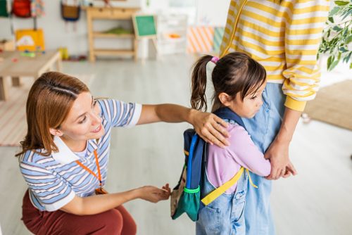 Preschool teacher in kindergarten classroom greeting new student, helping to easily assimilate with others.