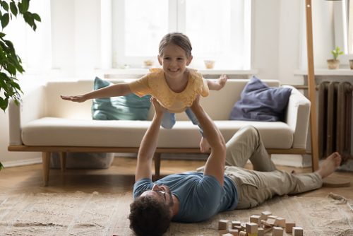 Flying with super dad. Caring joyful young father lying on warm floor in living room hold cheerful early school age daughter above chest on stretched arms play airplane pretend funny flight adventure