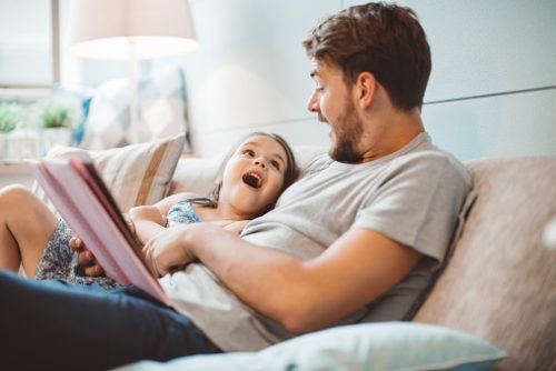 A father reads a bedtime story to his young daughter on the couch, both smiling and laughing together. Speech therapy in NYC supports children’s language and brain development through similar bonding activities.
