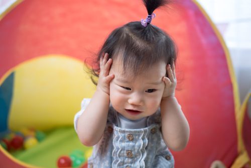 A toddler holding their head with both hands in a colorful play area, illustrating early frustration that can be addressed with pediatric speech therapy in NYC.